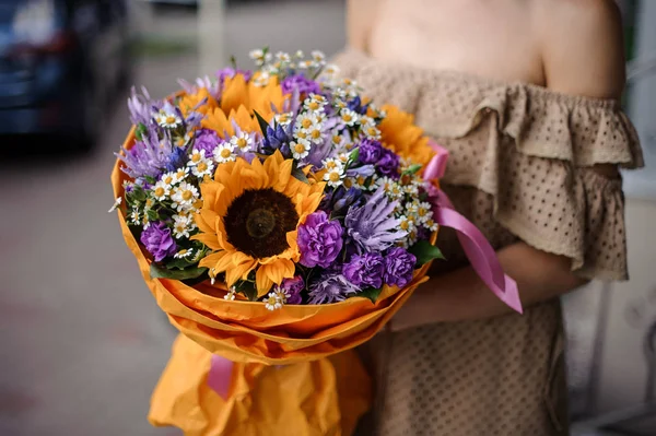 穿米色连衣裙的妇女抱着一束鲜艳的紫色花朵 向日葵和 Chamomiles 饰着蝴蝶结 — 图库照片