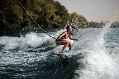Sexy smiling brunette woman wakesurfing on a board down the blue water on sunny day clipart