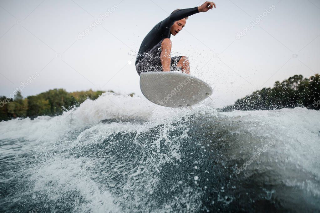 Active wakeboarder in swimsuit on the foreground jumping on the blue splashing wave against the background of clear grey sky