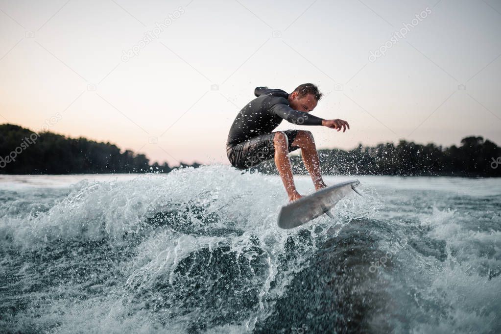 Professional wakeboarder in swimsuit jumping on the blue splashing wave against the background of clear grey sky on the foreground