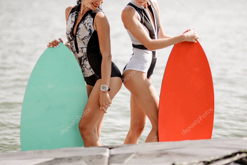 Two young sexy women dressed in black and white swimsuits standing with wakeboards against the water