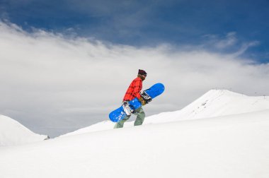 Kırmızı ekose desenli gömlek ve sıcak pantolon giyen mavi snowboard gökyüzüne karşı tutarak karla kaplı dağa, yükselir