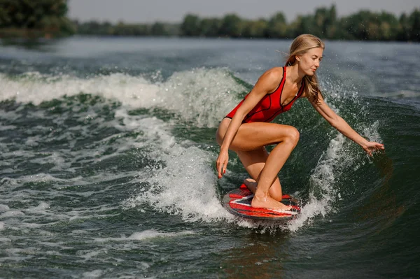 美丽的金发女孩在红色的泳装骑在红色的滑水在弯曲的膝盖和采取波浪与手 — 图库照片