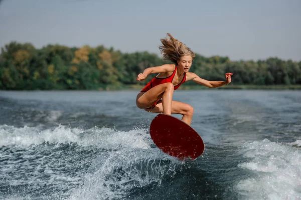 曲げ膝と取る手波赤ウェイク ボード上でジャンプ赤水着で美しいブロンドの女の子 — ストック写真