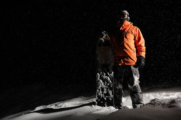 滑雪板在橙色的运动服和面具站立与董事会在一个下雪的山丘在夜间 — 图库照片