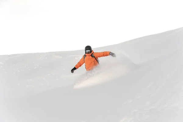 明るい白い空に対して粉の山の斜面の下の乗車オレンジのスポーツウェアのスノーボーダー — ストック写真