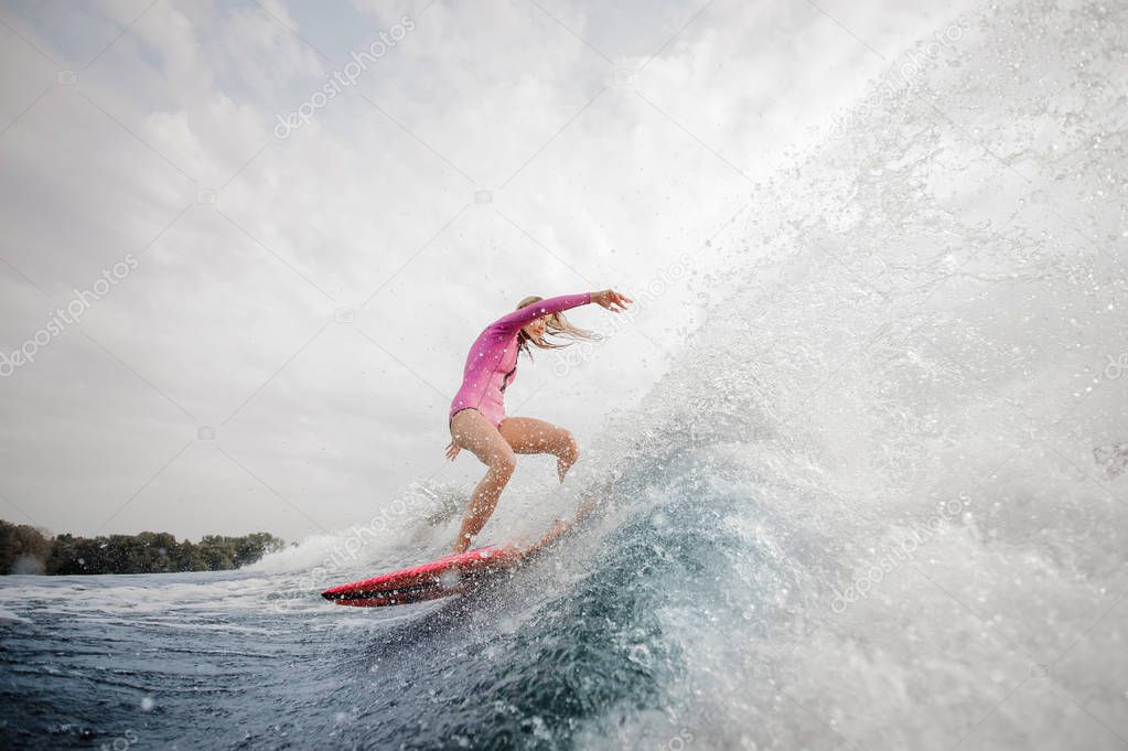 Active blonde woman wakesurfer dressed in pink swimsuit riding down the blue splashing wave against grey sky on a cloudy day