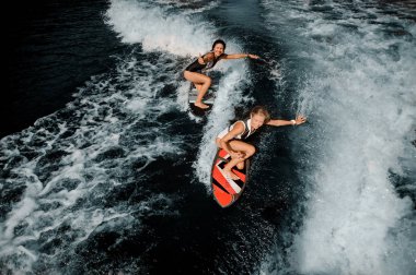 Two attractive girls in the black and white and red swimsuit riding on the wakeboards clipart