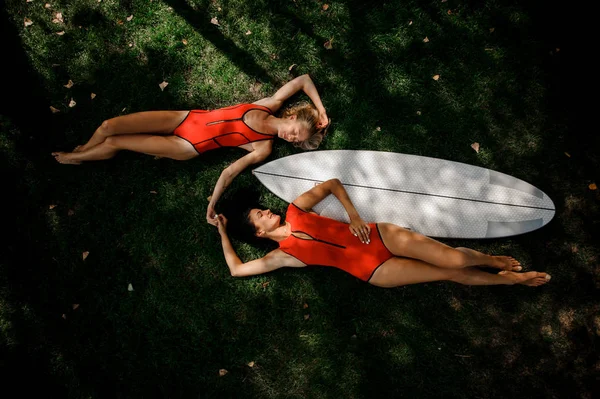 Two girls, blonde and brunette, in red swimsuits lying on the two sides of the white wakeboard on the green grass