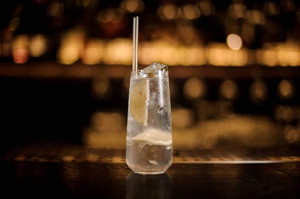 Long drink glass filled with Tom Collins cocktail on the bar counter against blurred golden lights