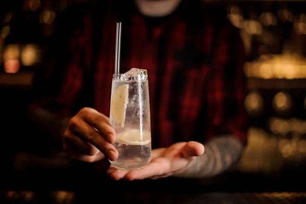 Bartender holding long drink glass filled with Tom Collins cocktail against the blurred background of lights