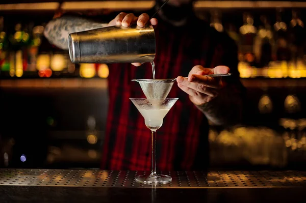 Bartender pourring a White Lady cocktail from the steel shaker through the sieve to a glass on the bar counter