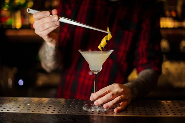 Bartender adding to a White Lady cocktail in the glass one orange zest with tweezers on the bar counter