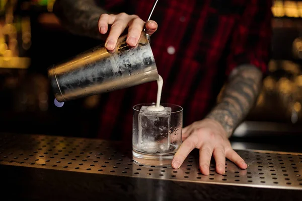 Barman pouring fresh creamy alcoholic drink into a glass with a big ice cube on bar