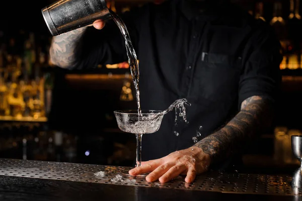 Professional bartender pouring an alcoholic drink from the steel shaker to the empty cocktail glass on the bar counter