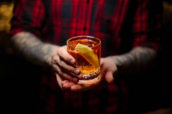 Barman tatuado con camisa sosteniendo una copa de cóctel con whisky — Foto de Stock