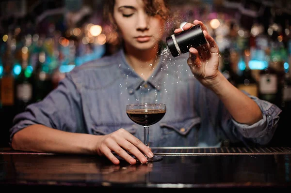 Chica camarera añadiendo a un cóctel con alcohol marrón un polvo en el mostrador de la barra —  Fotos de Stock