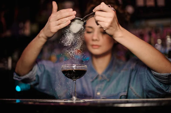 Bartender girl adding to a brown cocktail a powder through the strainer on the bar counter