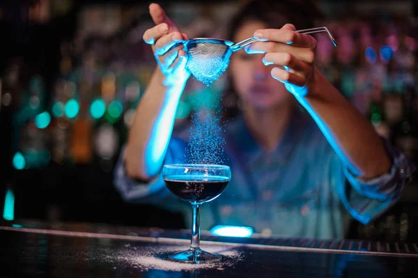 Female bartender adding to a brown cocktail a powder through the strainer in the blue light