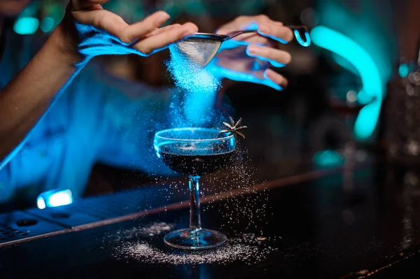 Female bartender adding to a cocktail decorated with badian a sugar powder through the strainer in the blue light