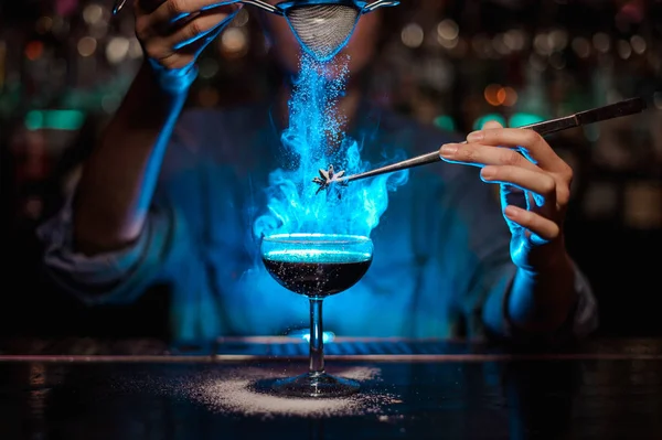 Chica camarera añadiendo a un cóctel marrón y verter en un badian flameado en pinzas de azúcar en polvo en la luz azul — Foto de Stock