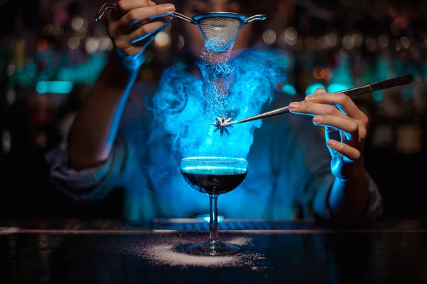 Cantinera hembra añadiendo a un cóctel marrón y verter en un badian flameado en pinzas un azúcar en polvo en la luz azul —  Fotos de Stock