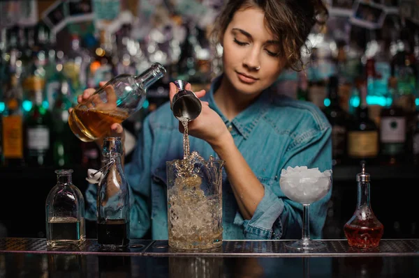 Female bartender pouring to the measuring glass cup with ice cubes an alcoholic drink from steel jigger — Stock Photo, Image