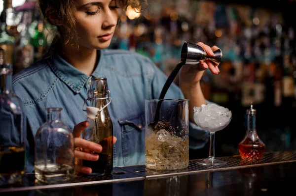 Barman feminino derramando para o copo de vidro de medição com cubos de gelo uma bebida alcoólica marrom de jigger de aço — Fotografia de Stock