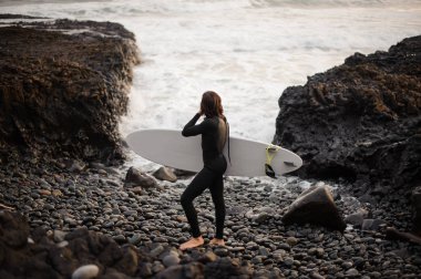 Arka görünümü adam elinde çakıl plajı okyanus arka planda kayaların arasındaki bir sörf ile ayakta.