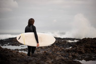 Arka görünümü esmer adam siyah mayoyla denize bakarak rock sahilde elinde bir sörf ile ayakta.