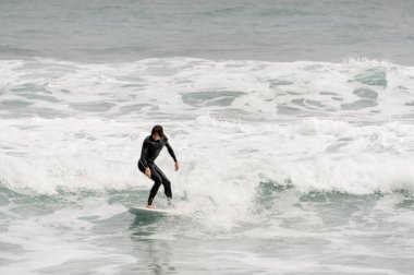 Esmer adam siyah mayo ayakta fırtınalı okyanus üzerinde beyaz bir sörf üzerinde.