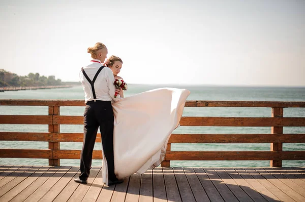Mladá a krásná manželský pár všeobjímající na dřevěný most, v pozadí moře — Stock fotografie