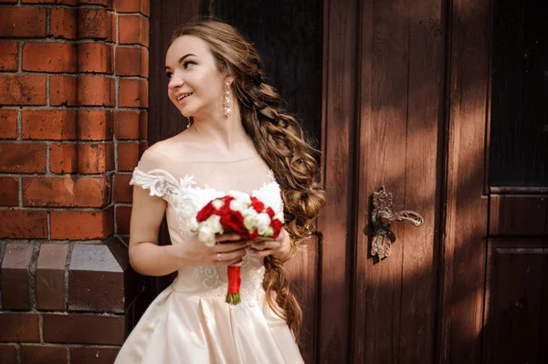 Hermosa novia en vestido de novia blanco con un ramo de boda de pie cerca de la puerta de madera — Foto de Stock