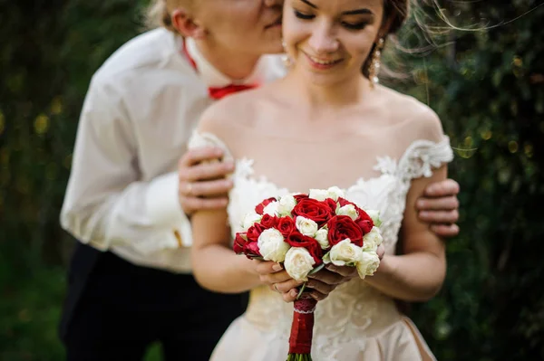 Groom kissing his young and beautiful bride in background of green tree — Stock Photo, Image