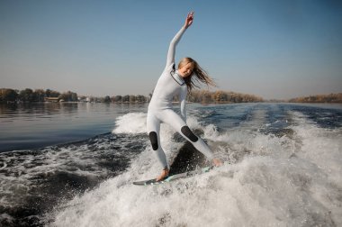 Happy beautiful blonde girl in the white swimsuit riding on the green wakeboard on the bending knees clipart