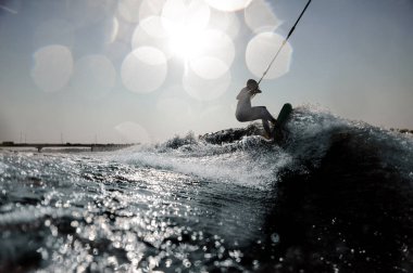 Beautiful blonde girl in the white swimsuit riding on the wakeboard holding a rope on the bending knees clipart