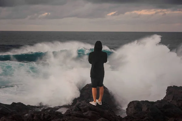 Jeune homme en sweat à capuche et short debout sur le rocher près de la mer orageuse — Photo