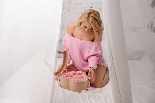 Menina loira na camisa rosa sentada na cama abrindo a caixa de flores olhando para ele — Fotografia de Stock