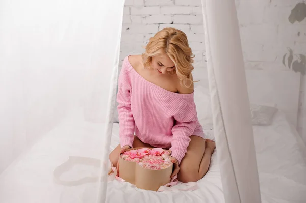 Chica con la camisa rosa sentada en la cama abriendo la caja de flores mirándola — Foto de Stock