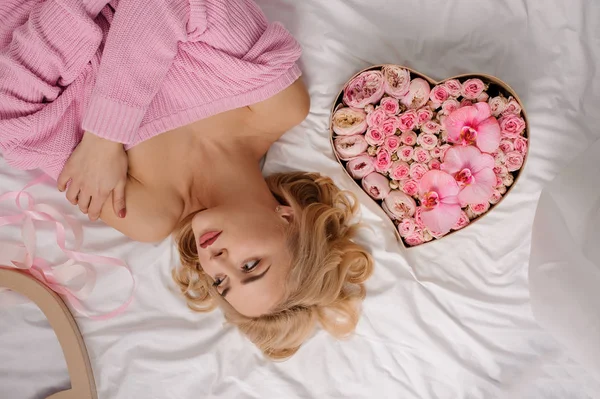 Krásná dívka v růžové košili leží na posteli poblíž tvaru srdce box barevné květy růže — Stock fotografie