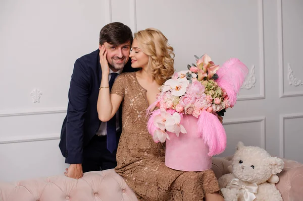 Fille dans la robe beige assis sur le canapé avec la boîte de fleurs étreignant son homme — Photo