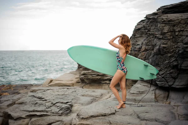 Vista lateral de uma menina bonita no maiô multi colorido de pé com o surf azul — Fotografia de Stock