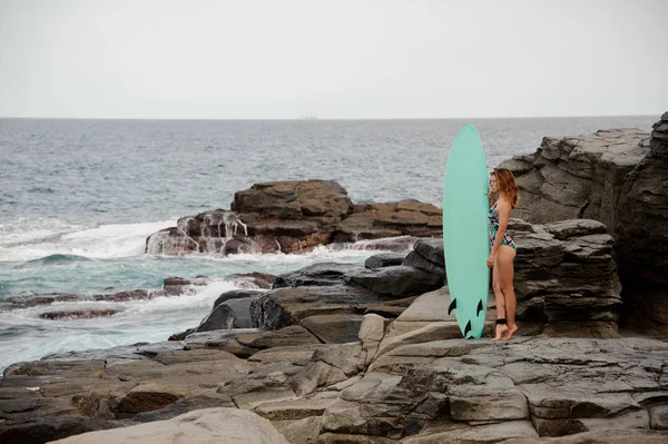 Belle fille dans le maillot de bain multicolore debout avec le surf sur la plage de roche — Photo