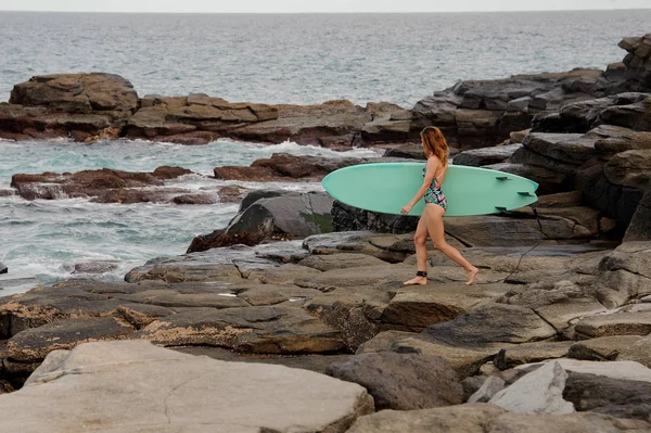 Menina desportiva no maiô multi colorido andando com o surf na praia de rock — Fotografia de Stock