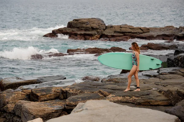 Fille sportive dans le maillot de bain multicolore marche avec le surf sur les rochers sur la plage — Photo