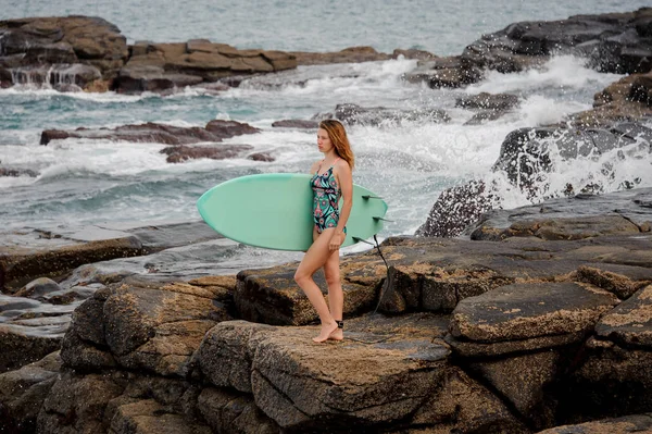 Modelo no maiô multicolorido andando com o surf nas rochas na praia — Fotografia de Stock