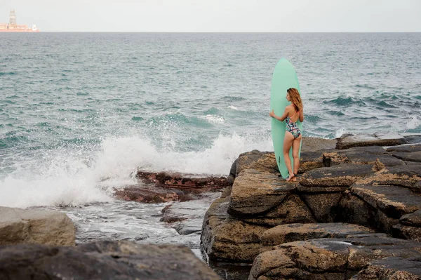 Menina ajuste fino no maiô multi colorido de pé com o surf nas rochas na praia — Fotografia de Stock