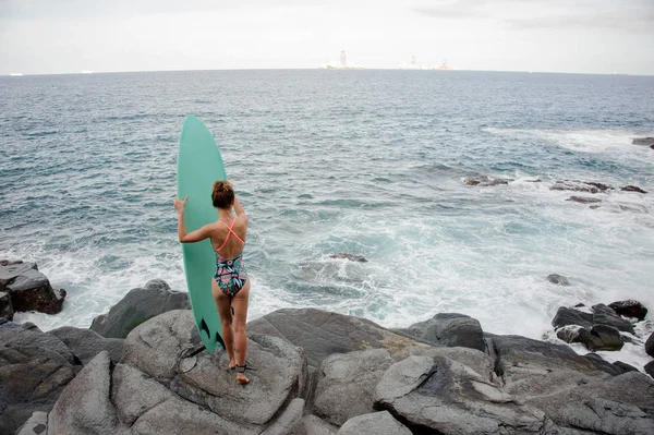 Vue arrière fille sexy dans le maillot de bain multicolore debout avec le surf sur la plage de roche — Photo