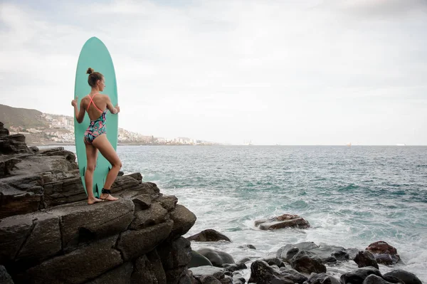 Menina morena no maiô multi colorido de pé com o surf na rocha — Fotografia de Stock