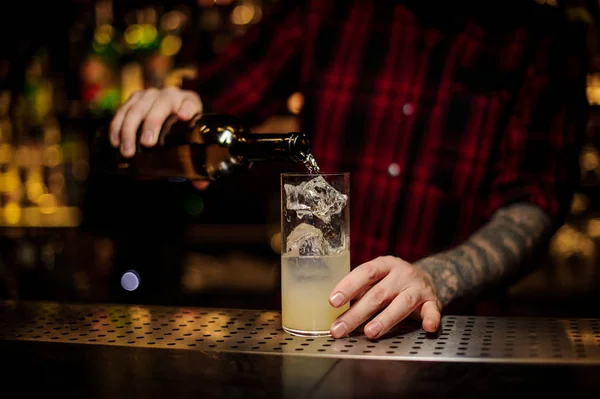 Mixologist pouring alcoholic drink from bottle into a cocktail glass with juice and ice cubes